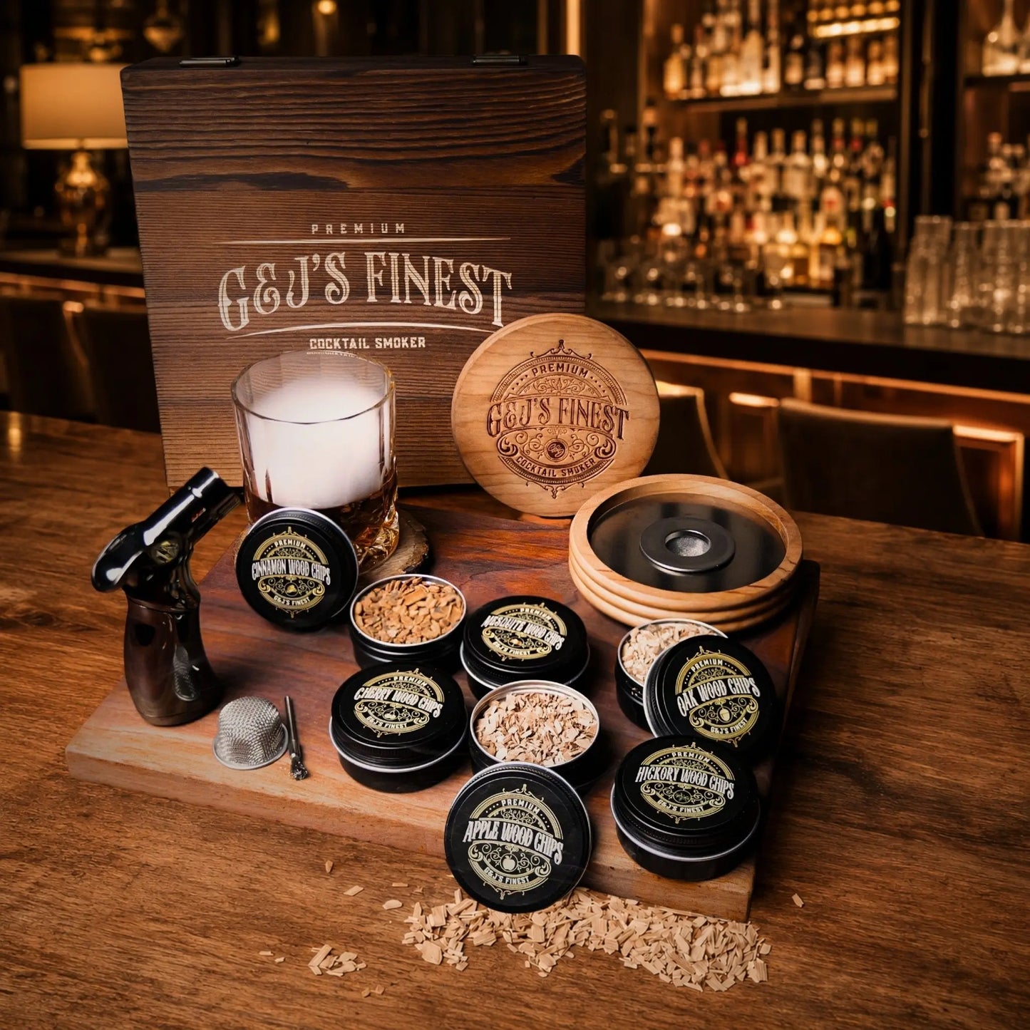 G&J's Finest Premium Cocktail Smoker Kit displaying a wooden gift box, a smoke-filled rocks glass with a round wood infuser lid, a black culinary torch, a metal mesh filter, and six tins of flavored wood chips including apple, oak, hickory, cherry, mesquite, and cinnamon on a wooden serving board. Cherry Wood Smoker