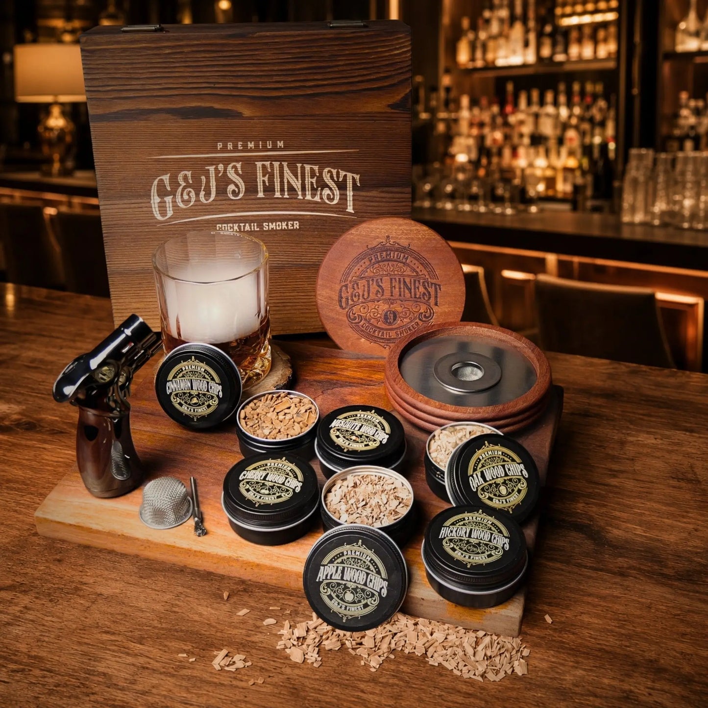 Whiskey smoker kit lineup with wood box, chip tins, glasses, and a torch on a dark bar tabletop.