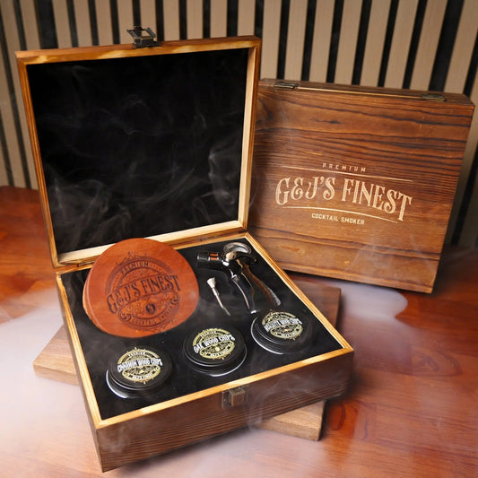 Open mahogany gift box showing a cocktail smoker top, glasses, and wood chips with smoke effects.